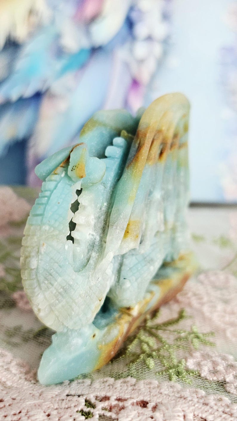 May include: A carved stone sculpture in shades of blue, tan, and white. The sculpture appears to be a stylized dragon or mythical creature. The details include carved scales and a textured surface. The sculpture is set against a soft, blurred background.