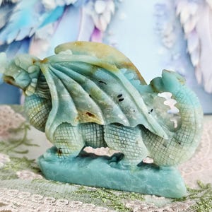 May include: A carved stone dragon sculpture in shades of teal, tan, and brown. The dragon has detailed wings, scales, and a curled tail. The sculpture is mounted on a base and is a decorative item.