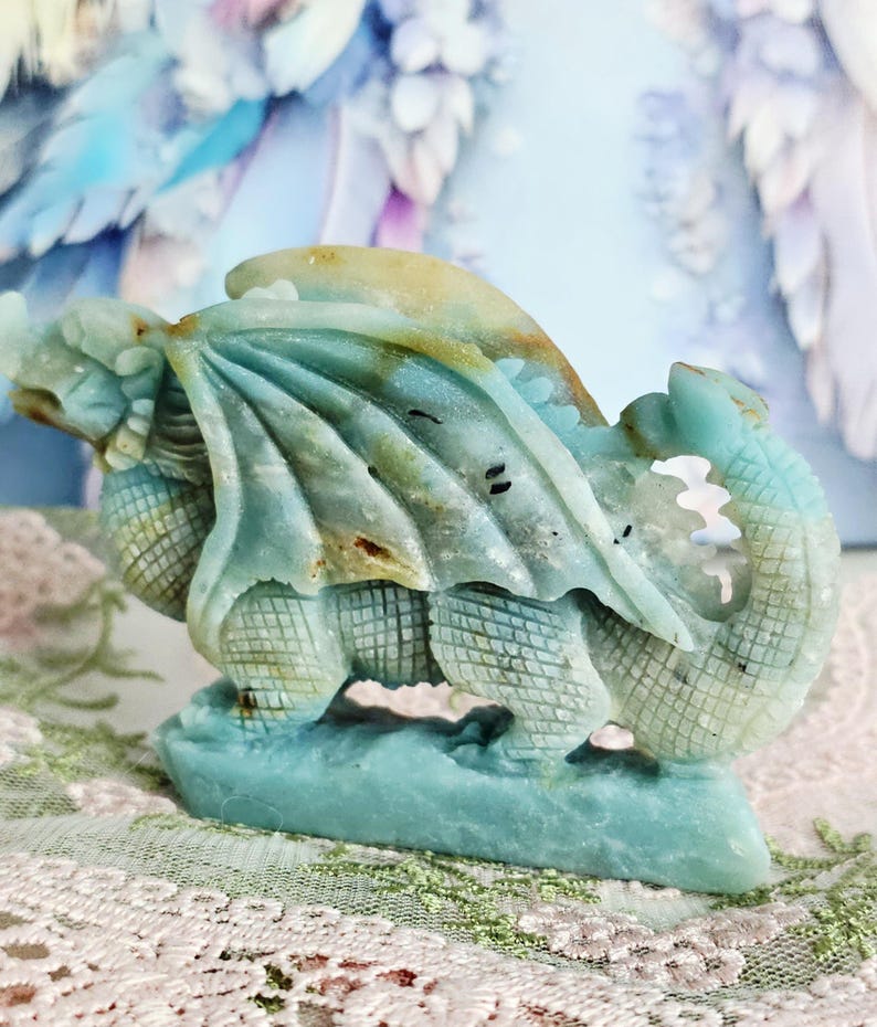 May include: A carved stone dragon sculpture in shades of teal, tan, and brown. The dragon has detailed wings, scales, and a curled tail. The sculpture is mounted on a base and is a decorative item.