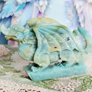 May include: A carved dragon figurine in shades of blue and tan. The dragon has detailed wings, scales, and a long tail. The figurine is made of a stone-like material and sits on a rectangular base. The dragon is a fantasy-themed decorative item.