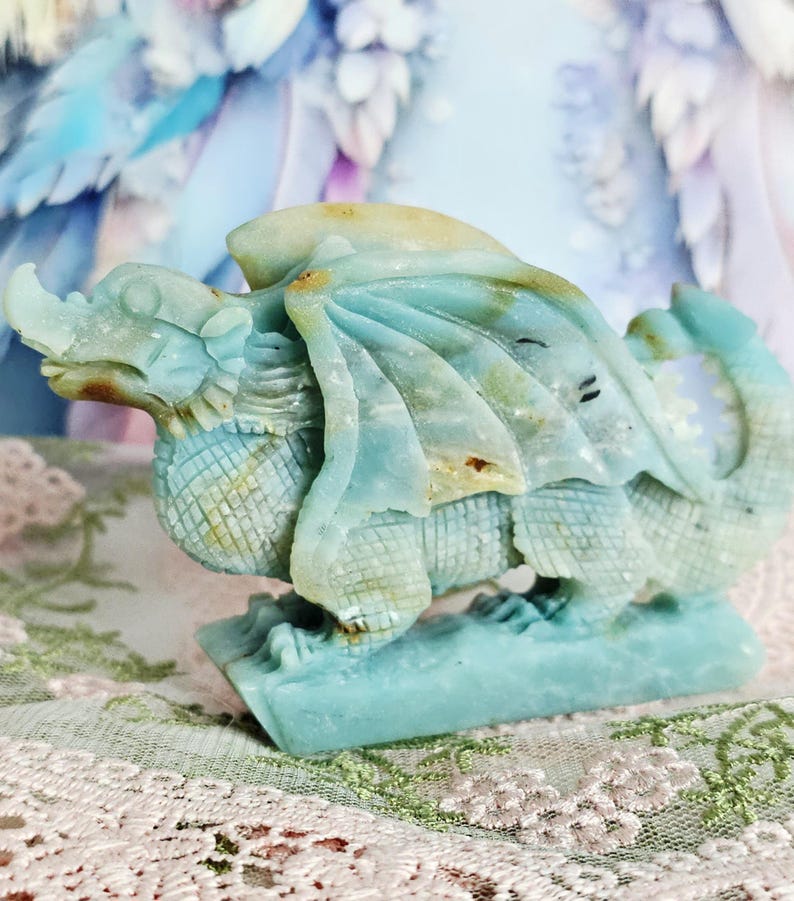 May include: A carved dragon figurine in shades of blue and tan. The dragon has detailed wings, scales, and a long tail. The figurine is made of a stone-like material and sits on a rectangular base. The dragon is a fantasy-themed decorative item.