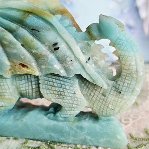 May include: A detailed sculpture of a dragon, carved from a stone with shades of blue, green, and tan. The dragon's scales and wings are intricately designed, with a textured appearance. The sculpture is set against a soft, blurred background.