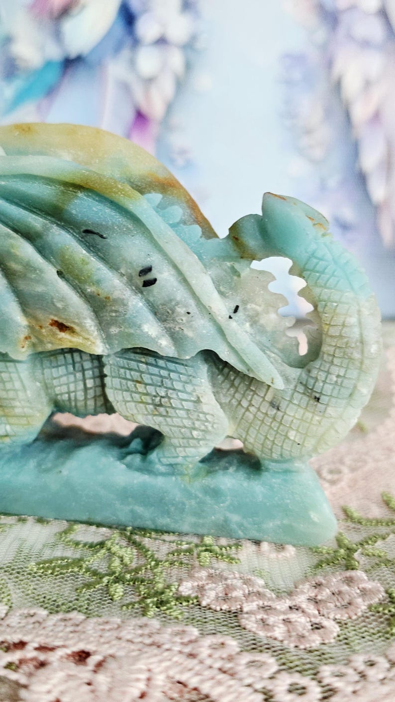 May include: A detailed sculpture of a dragon, carved from a stone with shades of blue, green, and tan. The dragon's scales and wings are intricately designed, with a textured appearance. The sculpture is set against a soft, blurred background.
