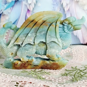 May include: A carved stone dragon figurine in shades of blue, green, and brown. The dragon has detailed wings, scales, and a textured base. The figurine is a decorative item, possibly for display or collection.