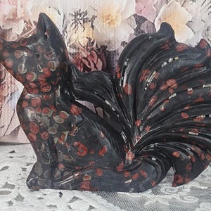 May include: A carved stone figurine of a fox with multiple tails, crafted from dark black stone featuring prominent circular red and grey spots throughout its body. The fox is depicted in a seated position with its large, bushy tails fanned out.