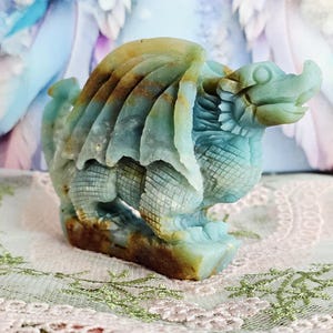 May include: A carved stone dragon figurine in shades of blue, brown, and tan. The dragon has detailed wings, scales, and a fierce expression. The figurine is standing on a rectangular base and is a decorative item.