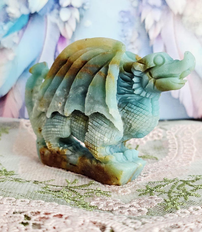 May include: A carved stone dragon figurine in shades of blue, brown, and tan. The dragon has detailed wings, scales, and a fierce expression. The figurine is standing on a rectangular base and is a decorative item.