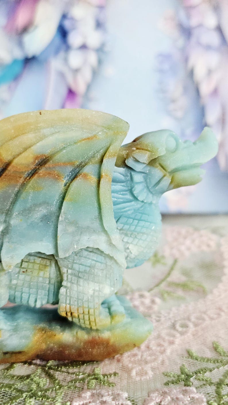 May include: A carved stone dragon figurine in shades of blue, green, and brown. The dragon has detailed wings, scales, and a textured body. The sculpture is a decorative item, possibly for display or collection.