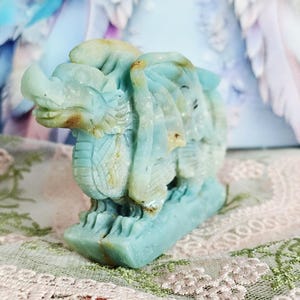 May include: A carved stone dragon figurine in shades of blue and tan. The dragon is detailed with scales, wings, and a fierce expression. The figurine is displayed on a decorative surface, showcasing the intricate craftsmanship of the sculpture.