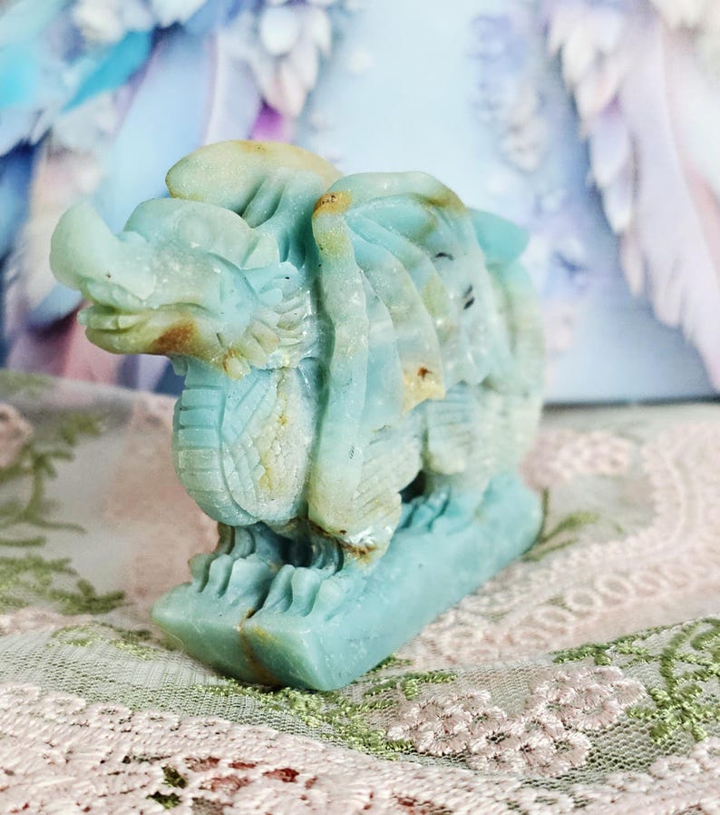 May include: A carved stone dragon figurine in shades of blue and tan. The dragon is detailed with scales, wings, and a fierce expression. The figurine is displayed on a decorative surface, showcasing the intricate craftsmanship of the sculpture.