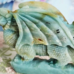 May include: A detailed, close-up of a turquoise dragon sculpture. The dragon has textured wings and scales, with hints of brown and yellow. The sculpture is set against a soft, blurred background.