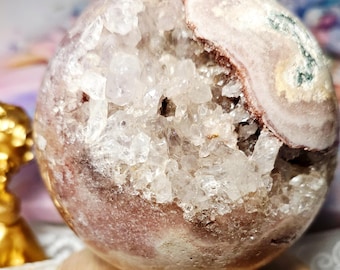 May include: A spherical, polished crystal geode with a cavity filled with clear quartz crystals. The exterior is a mix of pale pink, white, and brown hues. The geode sits on a small, round wooden stand.