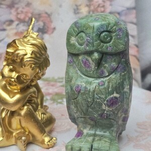 May include: A gold-colored figurine of a seated cherub with curly hair positioned next to a carved owl figurine made of green stone with distinct purple inclusions, featuring etched details on the face, wings, and feet of the owl.