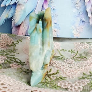 May include: A carved stone sculpture in shades of blue, white, and brown. The sculpture has a hand-like shape with multiple fingers. The background includes a light blue canvas with a floral design and a pink lace fabric.