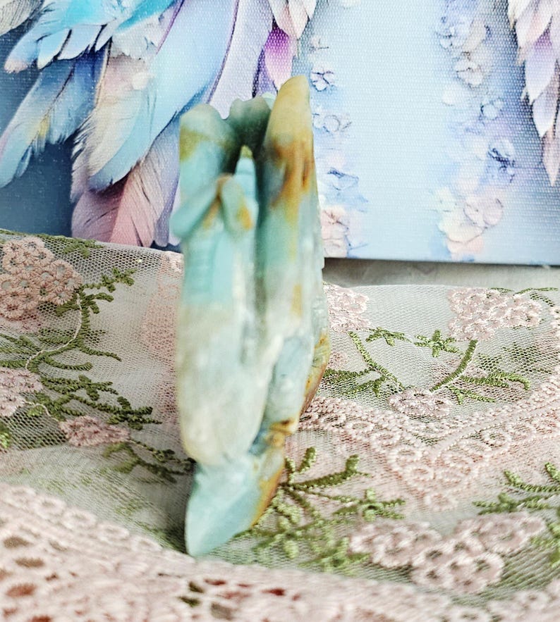May include: A carved stone sculpture in shades of blue, white, and brown. The sculpture has a hand-like shape with multiple fingers. The background includes a light blue canvas with a floral design and a pink lace fabric.