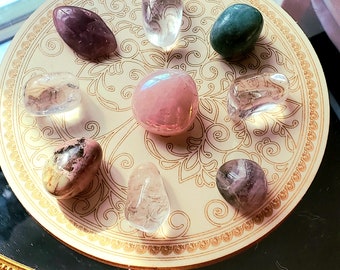 May include: A collection of seven tumbled stones, including clear quartz, rose quartz, amethyst, green aventurine, and a pink and white striped stone, arranged on a decorative plate with a floral pattern.