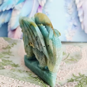 May include: A carved stone dragon figurine in shades of blue, green, and tan. The dragon is intricately detailed with wings and scales. The figurine is displayed on a lace-covered surface.