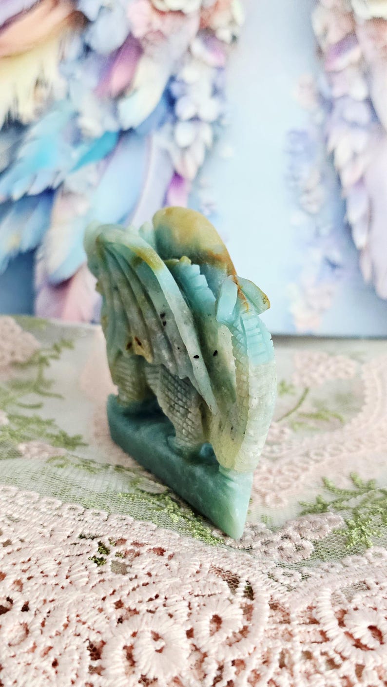 May include: A carved stone dragon figurine in shades of blue, green, and tan. The dragon is intricately detailed with wings and scales. The figurine is displayed on a lace-covered surface.