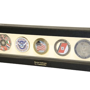 Op de afbeelding: Een zwarte rechthoekige displaykast met een glazen voorzijde die vijf verschillende challenge-munten toont. De munten tonen verschillende emblemen, waaronder het zegel van het Amerikaanse Department of Homeland Security, een Amerikaanse vlag en een embleem van de Amerikaanse kustwacht. Een naamplaatje onder de munten luidt "Steven Anderson, Lt. Commander."