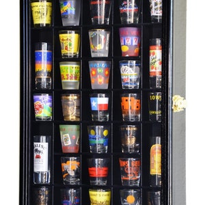 May include: Black wooden shot glass display case with glass shelves and a glass door. The case is filled with shot glasses featuring various designs and locations printed on them.