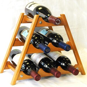 May include: A wooden wine rack with a triangular shape holds six bottles of wine. The rack is made of light brown wood and has a natural finish. The bottles are all red wine and are arranged in two rows of three. The bottles are all different brands and have different labels.
