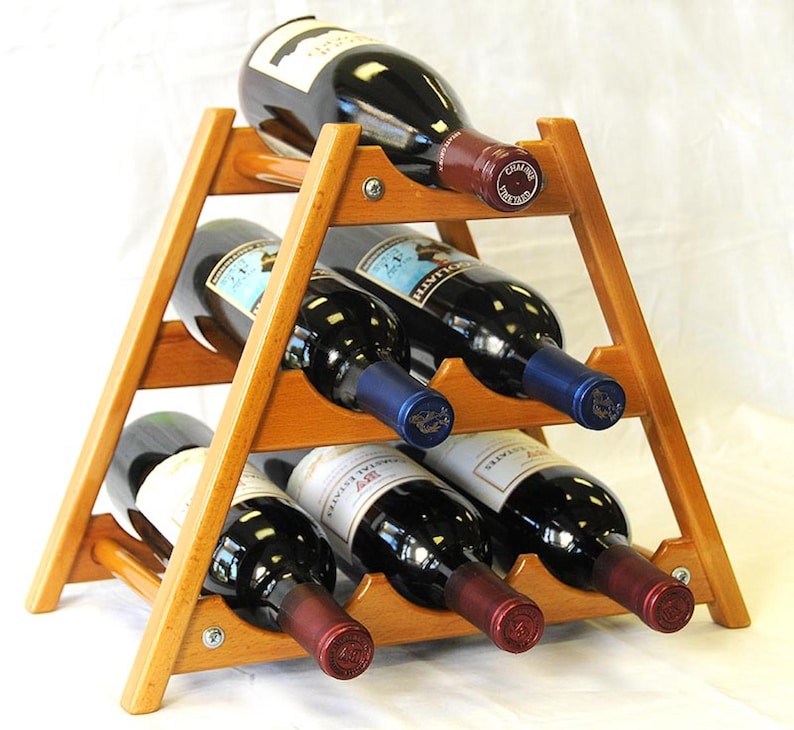May include: A wooden wine rack with a triangular shape holds six bottles of wine. The rack is made of light brown wood and has a natural finish. The bottles are all red wine and are arranged in two rows of three. The bottles are all different brands and have different labels.