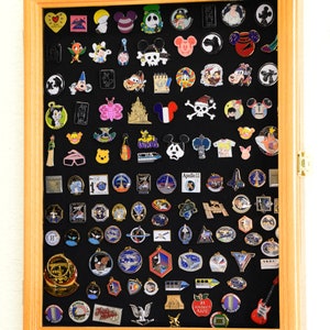 May include: A wooden shadow box display case with a black felt background filled with a variety of colorful enamel pins. The pins feature various themes including Disney characters, space exploration, and transportation.