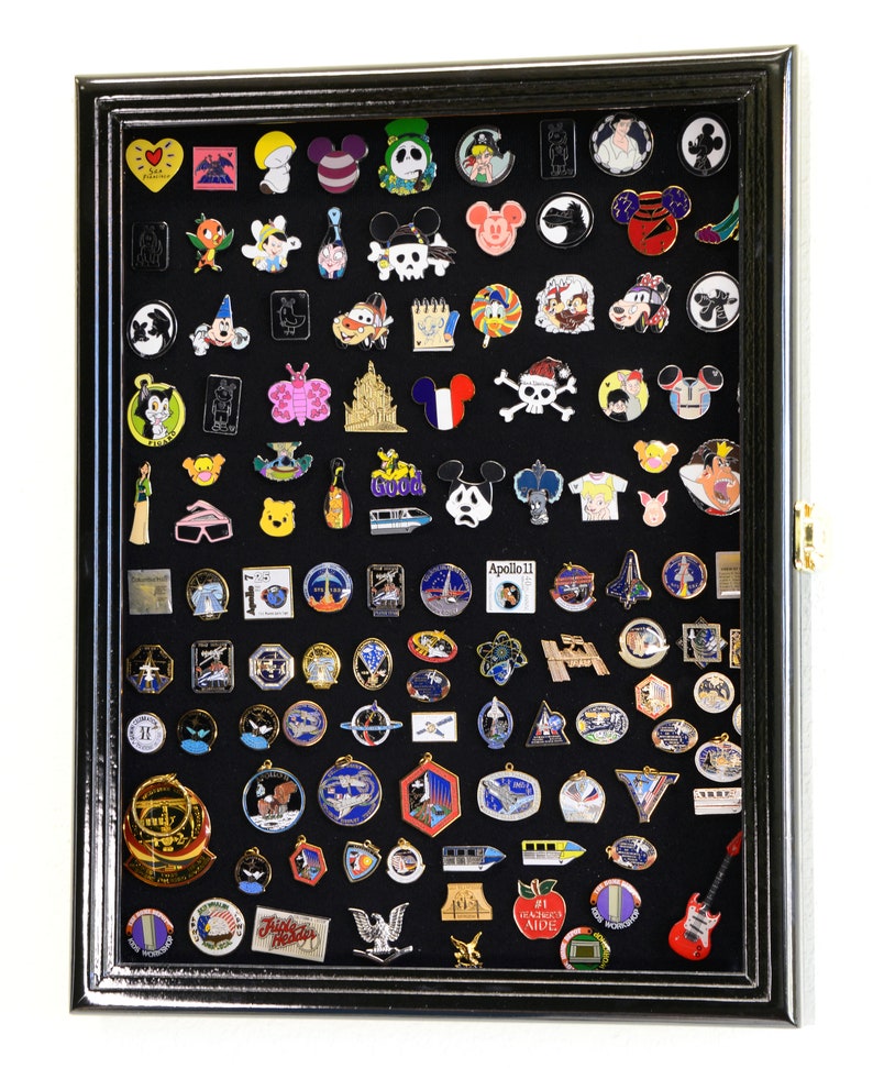 May include: A black shadow box display case with a black felt background filled with a variety of colorful enamel pins. The pins feature cartoon characters, logos, and other designs. The case has a glass front and a black frame.