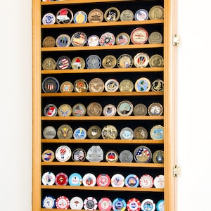 May include: A wooden display case with black velvet lining, holding a collection of challenge coins and casino chips. The case has a hinged door and is made of light wood.