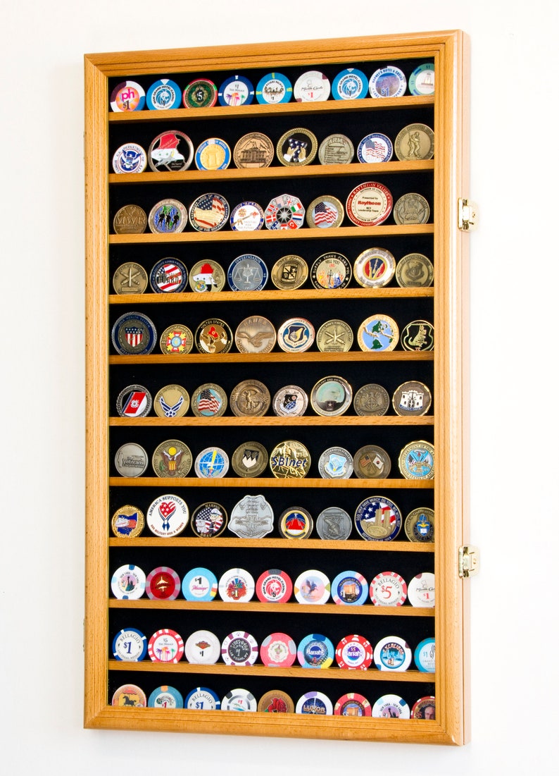 May include: A wooden display case with black velvet lining, holding a collection of challenge coins and casino chips. The case has a hinged door and is made of light wood.