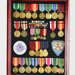 May include: A mahogany wood shadow box display case with a black felt background. The case is filled with military medals, ribbons, and patches. The medals are gold and silver and feature various designs, including eagles, stars, and anchors. The ribbons are red, white, blue, and green. The patches are embroidered with the words "The Essential Link" and "NAF" and feature a plane and a cross.