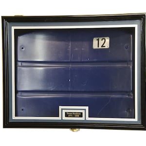 May include: A framed piece of blue stadium seating with a silver plaque that reads "12". The plaque is attached to the seating with two screws. The frame is black with a silver inner border. A small plaque at the bottom of the frame reads "Texas Stadium 1971-2008".