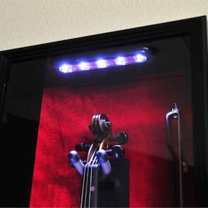 May include: A black wooden display case with a glass front and a red velvet interior. The case is illuminated by a row of white LED lights and features a violin inside.