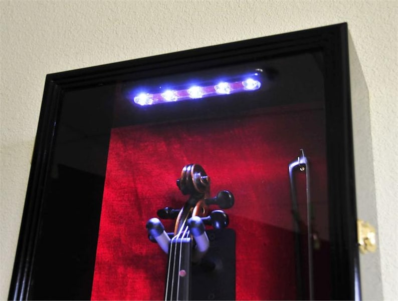 May include: A black wooden display case with a glass front and a red velvet interior. The case is illuminated by a row of white LED lights and features a violin inside.