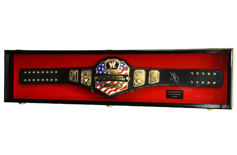 May include: A black leather wrestling championship belt with gold accents and a red, white, and blue center plate. The belt is displayed in a black and red display case. The center plate features the text "United States Champion" and a stylized "W".
