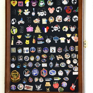 May include: A wooden shadow box display case with a black felt background filled with a variety of enamel pins. The pins feature a variety of themes including Disney characters, space exploration, and other popular icons.