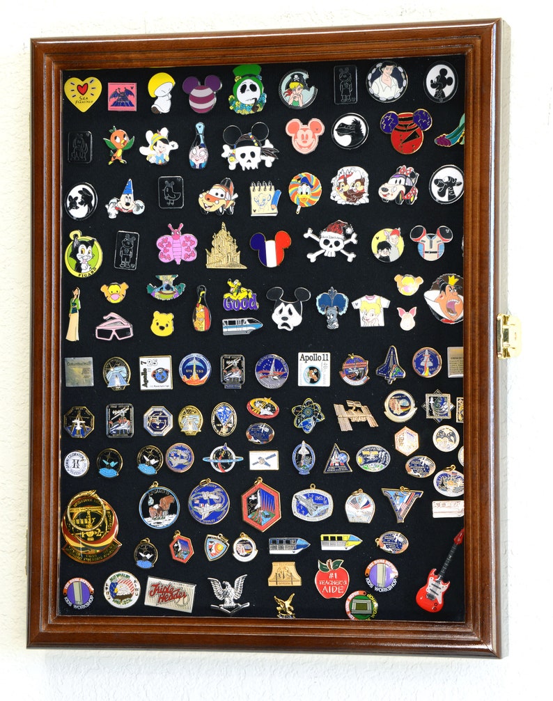 May include: A wooden shadow box display case with a black felt background filled with a variety of enamel pins. The pins feature a variety of themes including Disney characters, space exploration, and other popular icons.
