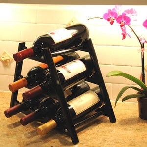 May include: Black metal wine rack with six bottles of wine. The rack is on a countertop with a light brown surface. A pink orchid plant is in a brown pot on the right side of the image.