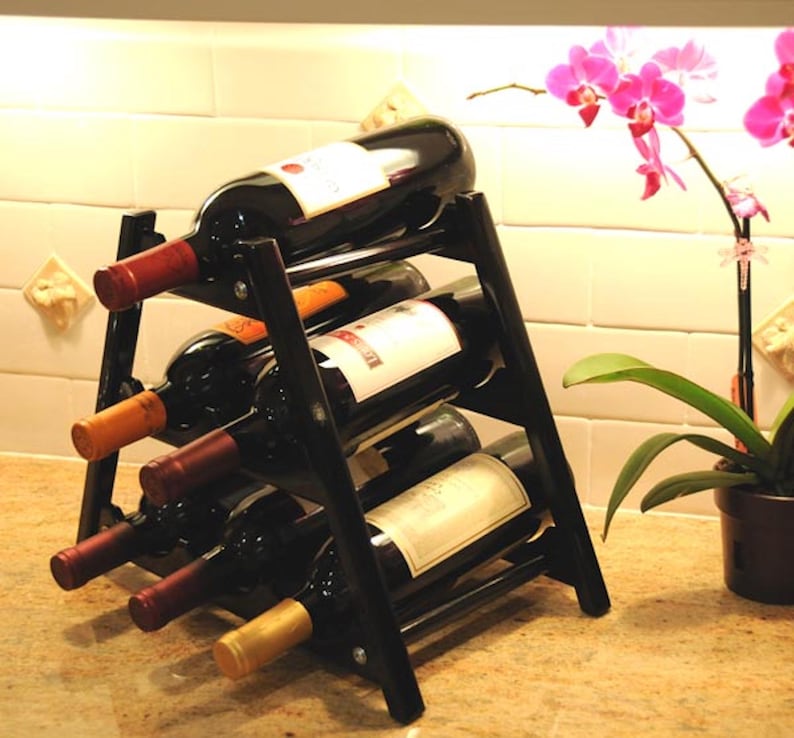 May include: Black metal wine rack with six bottles of wine. The rack is on a countertop with a light brown surface. A pink orchid plant is in a brown pot on the right side of the image.