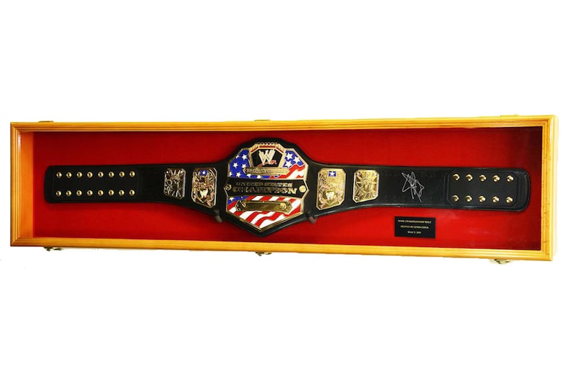 May include: A black leather wrestling championship belt with gold accents and a red, white, and blue American flag design. The belt is displayed in a wooden shadow box with a red velvet lining. The belt is signed by the wrestler.