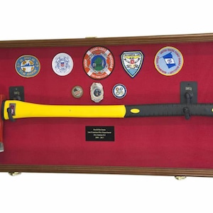 Pu&ograve; includere: Una scatola di ombra in legno con un rivestimento in feltro rosso mostra un'ascia da pompiere con un manico giallo e una testa nera. La scatola presenta anche diversi distintivi e spille, tra cui un emblema della Guardia Costiera e una toppa della Guardia Costiera Ausiliaria degli Stati Uniti.