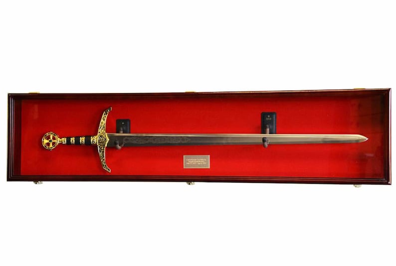 May include: A decorative sword in a wooden display case. The sword features a gold cross-shaped hilt and a detailed blade. The display case has a red velvet interior and a gold plaque. The sword is mounted on black supports.