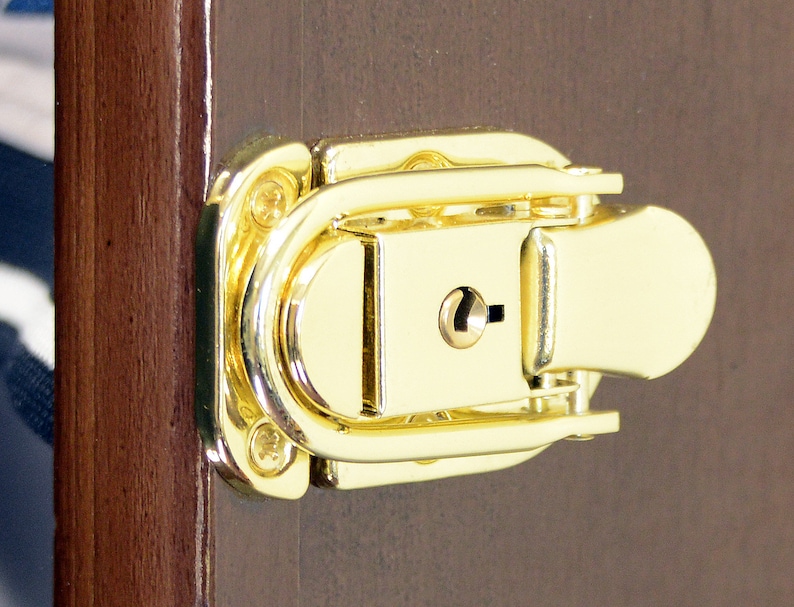 May include: A gold-toned metal latch with a keyhole. The latch is attached to a wooden surface.