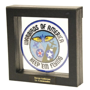 May include: A black shadow box with a round embroidered patch inside. The patch is blue with white lettering that reads "Warbirds of America Keep 'em Flying". A gold nameplate at the bottom reads "Steven Anderson Lt. Commander".