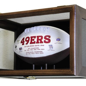Könnte beinhalten: Ein braunes Holzdisplay mit einer Glasfront und einem schwarzen Samt-Innenraum. Ein weißer Fußball mit roter Aufschrift "49ERS" ist im Display ausgestellt. Der Fußball hat zusätzlichen Text, der "ESTABLISHED DATE: 1946", "NFC CHAMPIONS", "1981, 1984, 1988, 1989, 1994, 2012", "SUPER BOWL CHAMPIONS", "XIX, XXIII, XXIV, XXIX" lautet.