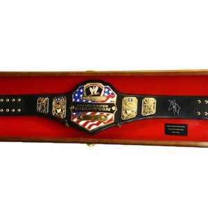 May include: A black leather wrestling championship belt with gold accents and a red, white, and blue center plate. The belt is displayed in a wooden shadow box with a red velvet lining. The center plate features the text "United States Championship".