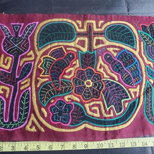 Puede incluir: Una obra de arte textil bordada de colores con fondo granate. El dise&ntilde;o presenta una flor central con un centro negro y un contorno amarillo, rodeada de mariposas, hojas y flores. La pieza est&aacute; enmarcada por un borde dorado.