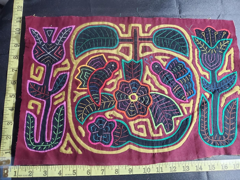 Puede incluir: Una obra de arte textil bordada de colores con fondo granate. El dise&ntilde;o presenta una flor central con un centro negro y un contorno amarillo, rodeada de mariposas, hojas y flores. La pieza est&aacute; enmarcada por un borde dorado.