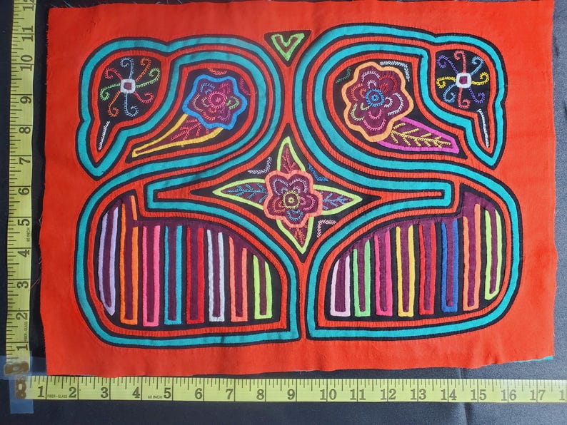 Puede incluir: Un textil mola colorido con un dise&ntilde;o geom&eacute;trico que presenta un motivo floral central y dos p&aacute;jaros a cada lado. El mola est&aacute; hecho de tela y est&aacute; cosido con hilos de colores.