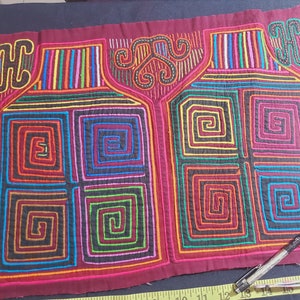 Puede incluir: Un tejido mola colorido con fondo rojo y dise&ntilde;os geom&eacute;tricos intrincados en tonos de azul, verde, amarillo, naranja y rosa. La mola presenta dos patrones sim&eacute;tricos de cuadrados conc&eacute;ntricos y espirales.